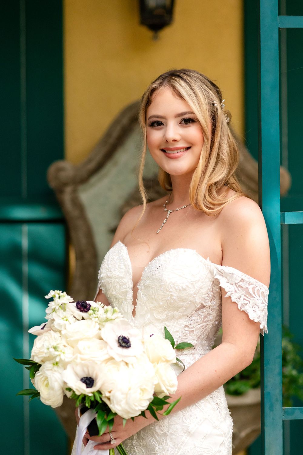 A bride in an off-shoulder lace wedding dress holds a white floral bouquet while smiling.