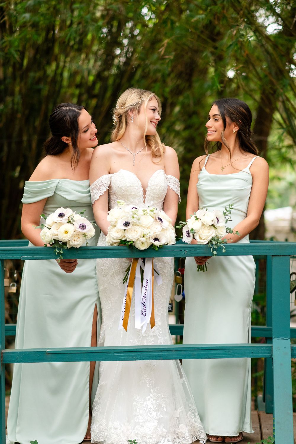 Bridesmaids in mint green dresses lean on a teal railing while holding white bouquets during an outdoor wedding.