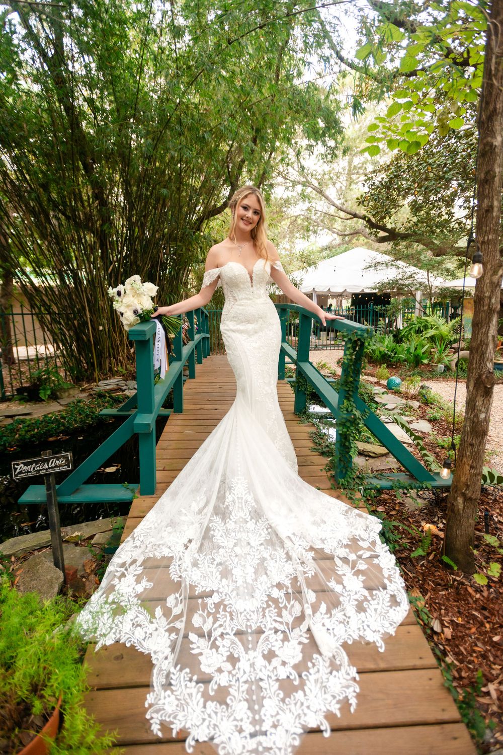 Flowing white lace wedding dress twirls on wooden boardwalk surrounded by green foliage.