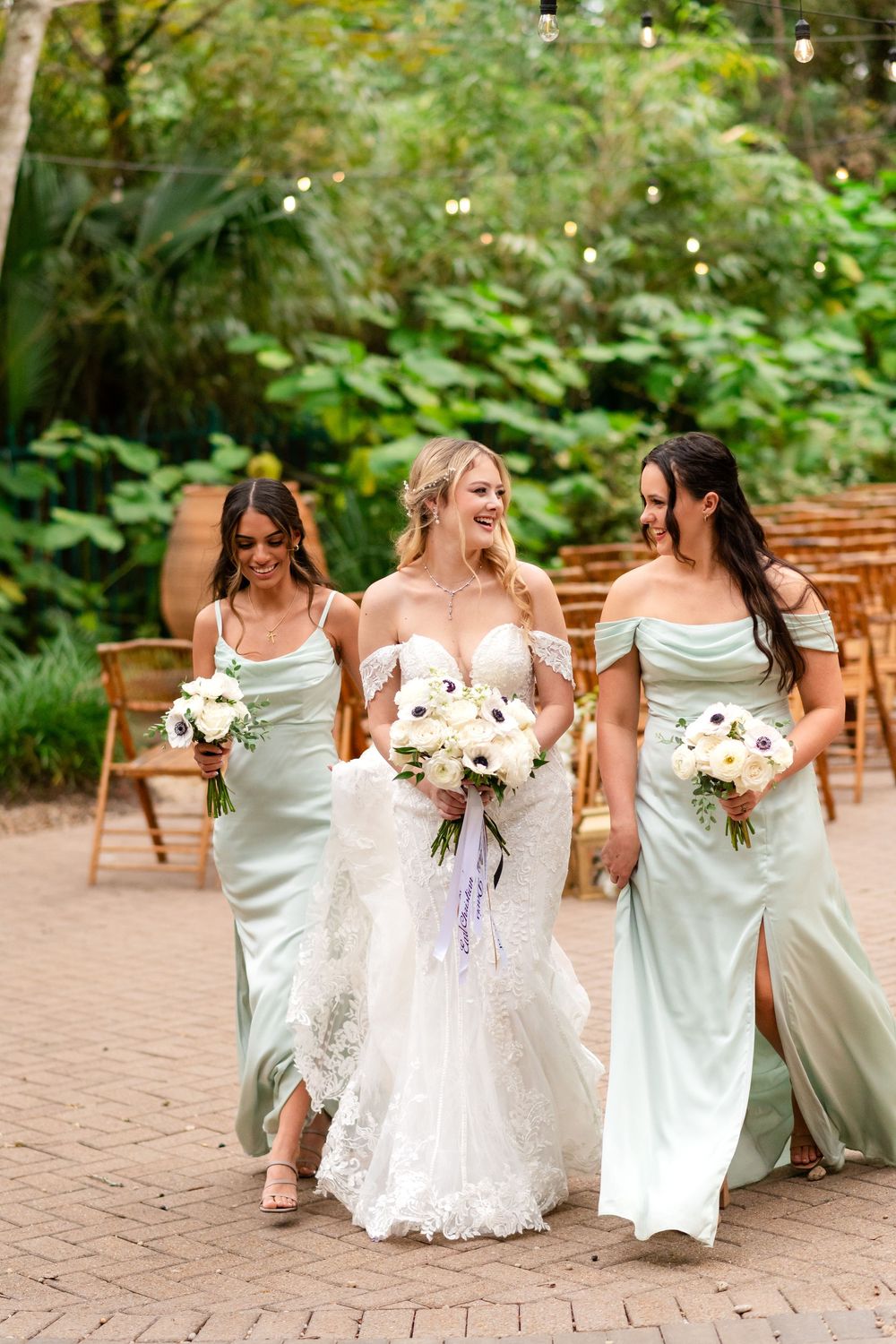 A bridal party in white dresses shares joyful moments together on a brick patio surrounded by lush greenery.