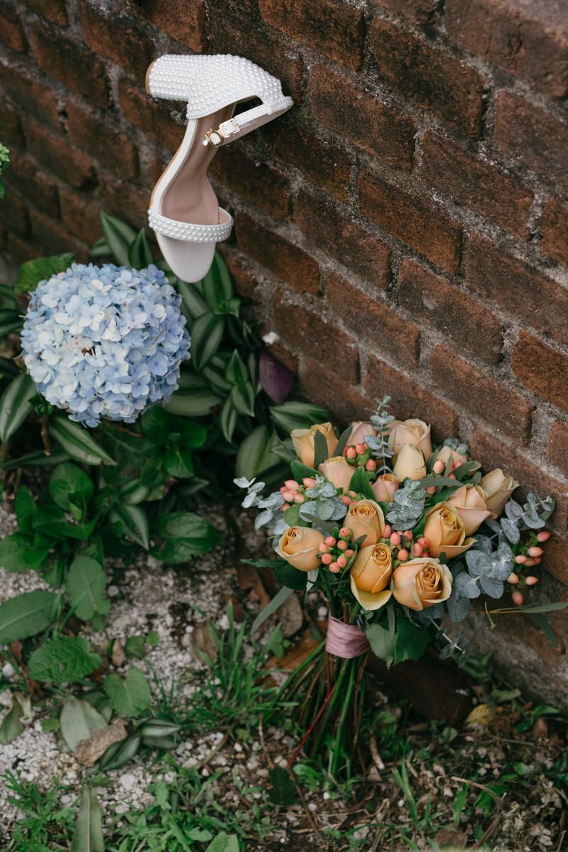 Wedding bouquet with peach roses and blue hydrangeas next to white bridal shoes against a brick wall.