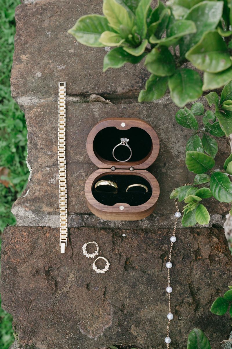 Wedding rings and jewelry displayed on rustic stone surface with green foliage, featuring engagement ring in wooden box.