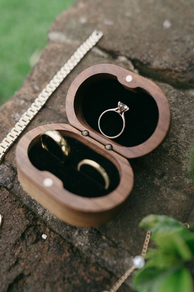 Wedding rings nestled in a heart-shaped wooden box displayed on rustic stone surface with green foliage in background.