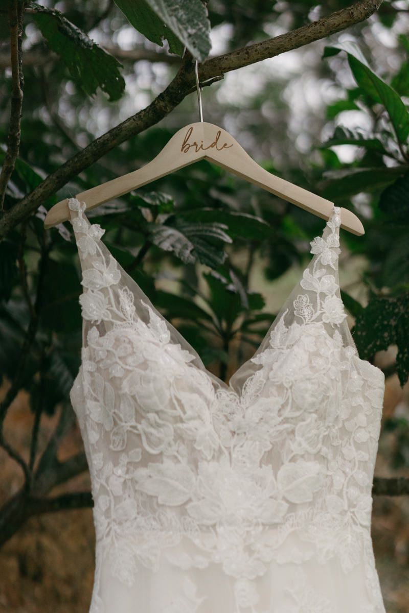 Close-up detail of a white lace wedding gown bodice with intricate floral patterns hanging on a wooden hanger.
