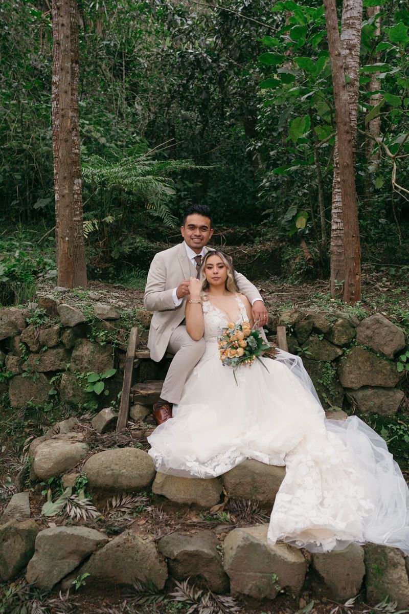 Couple poses on rustic stone wall surrounded by forest greenery.