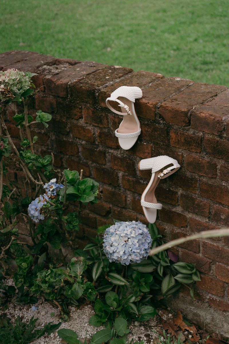 White bridal sandals displayed on a rustic brick wall with blue hydrangea flowers.