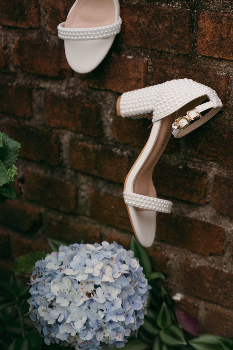 Pearl-embellished white wedding sandal displayed against a brick wall with a blue hydrangea bloom.