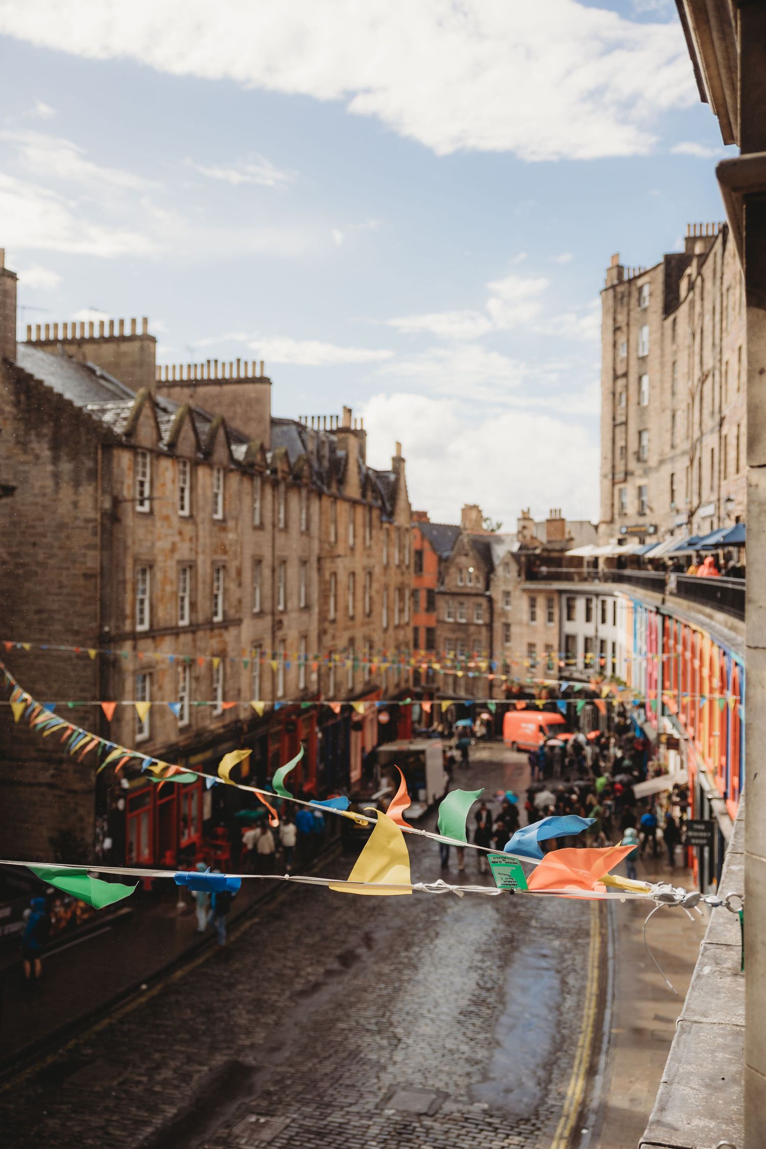Colorful bunting hangs over a wet cobblestone street lined with historic stone buildings in Edinburgh's old town.