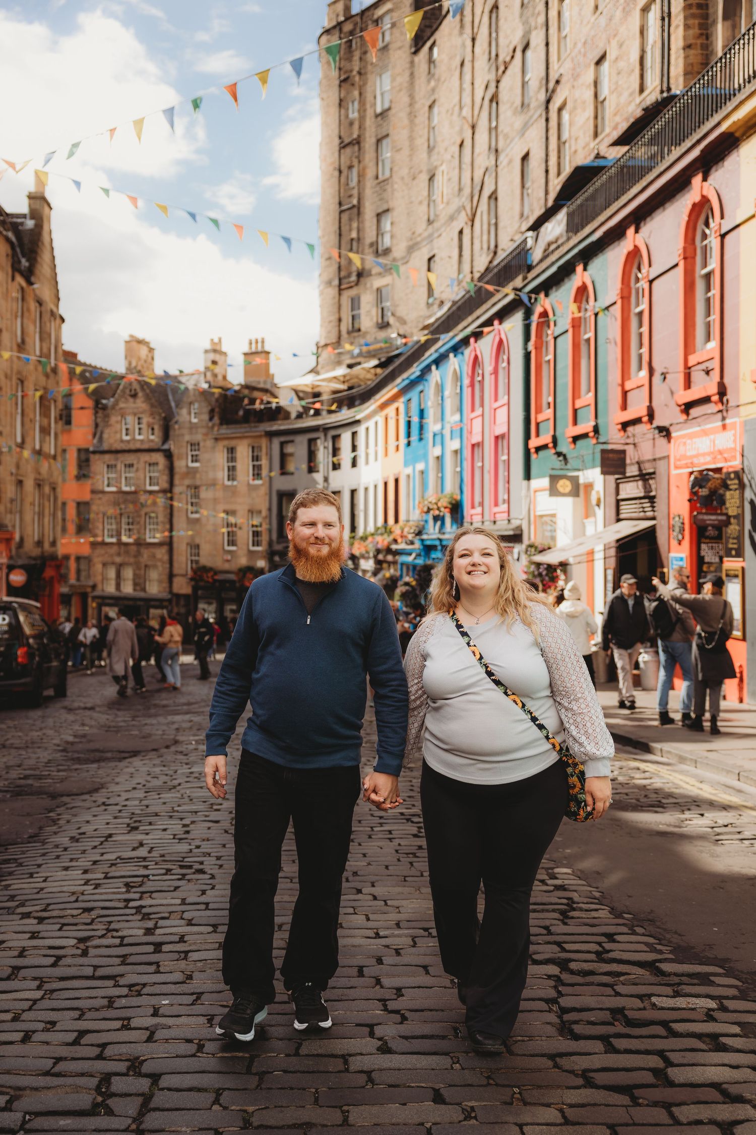 A couple walks together on a historic cobblestone street in Edinburgh with colorful buildings and bunting in the background.