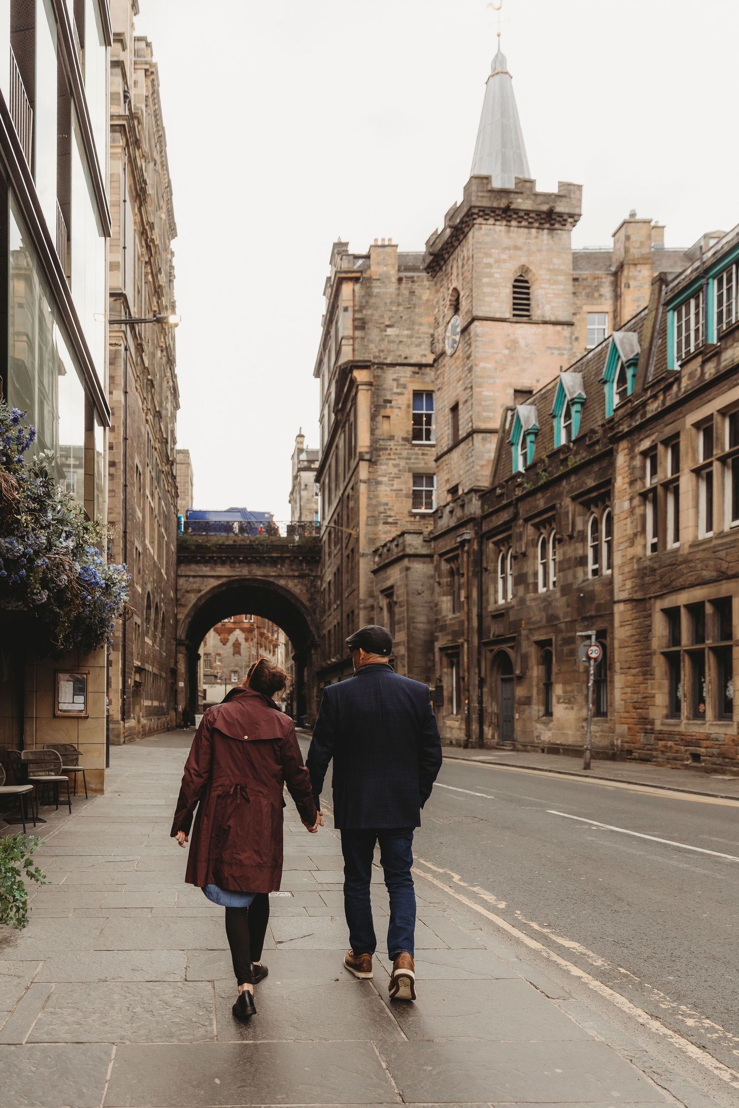 A couple walks down a historic cobblestone street in Edinburgh with gothic architecture and stone buildings in view.