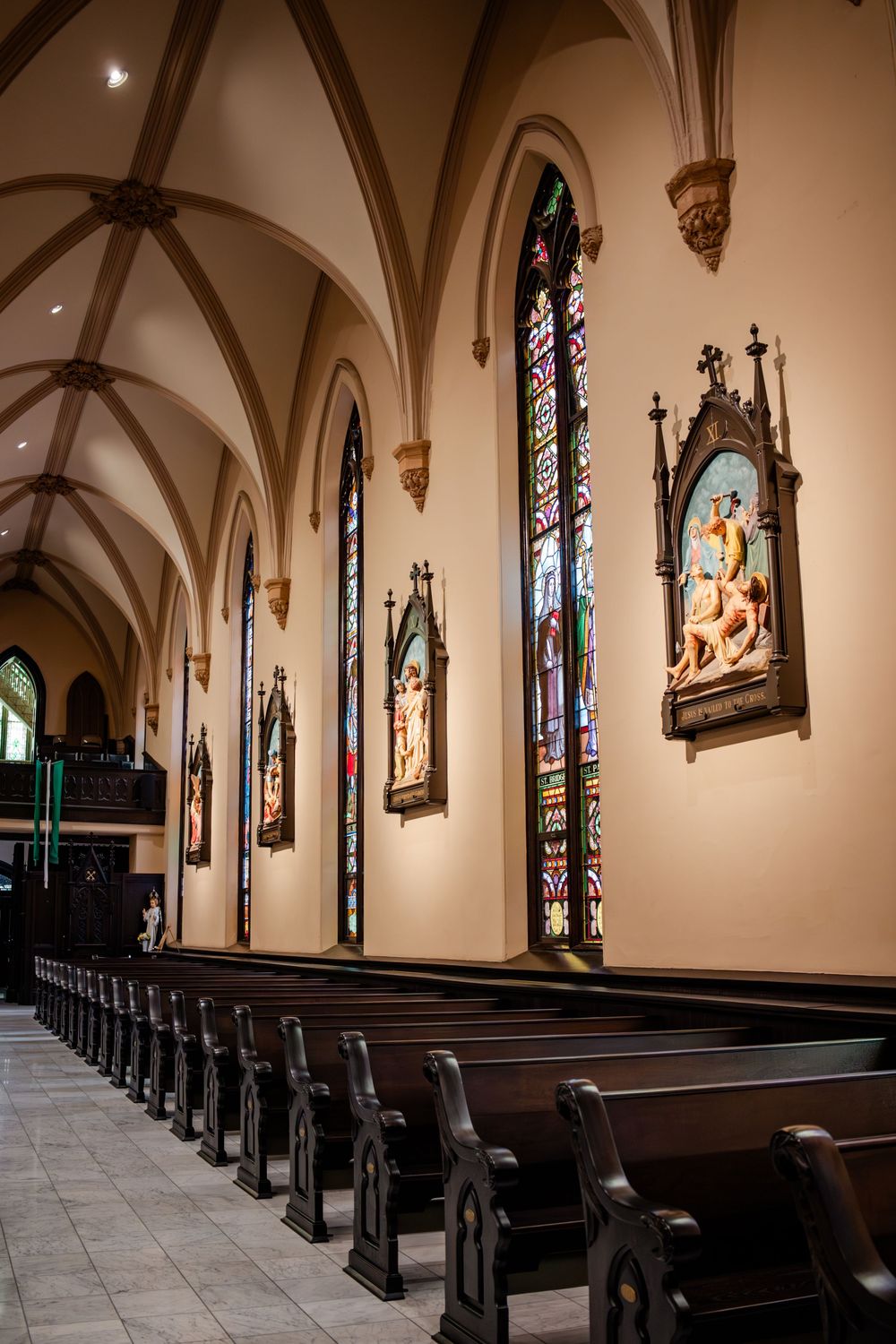 Gothic church interior with stained glass windows, wooden pews and religious artwork on vaulted walls.