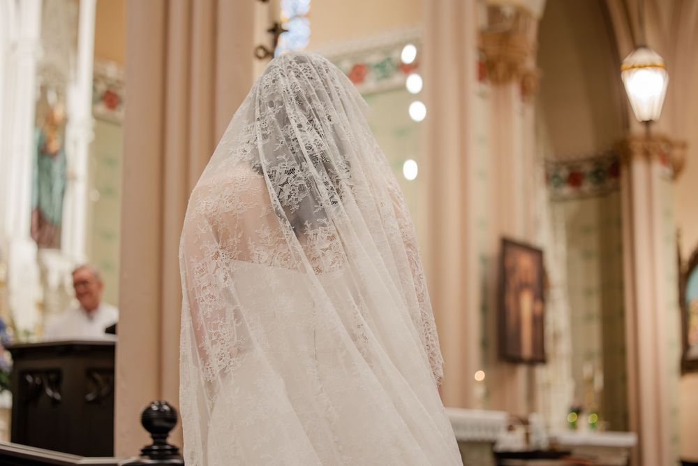 A bride in a lace veil stands in an elegant church setting with ornate decor and soft lighting.
