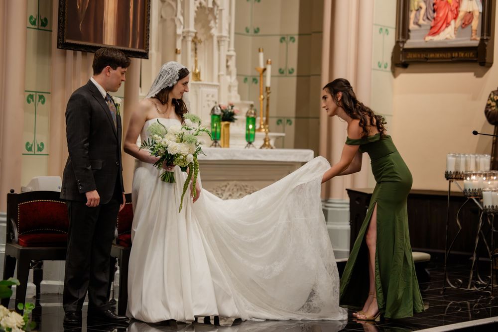 Wedding party members in formal attire arrange the bride's flowing white dress train inside an elegant church setting.