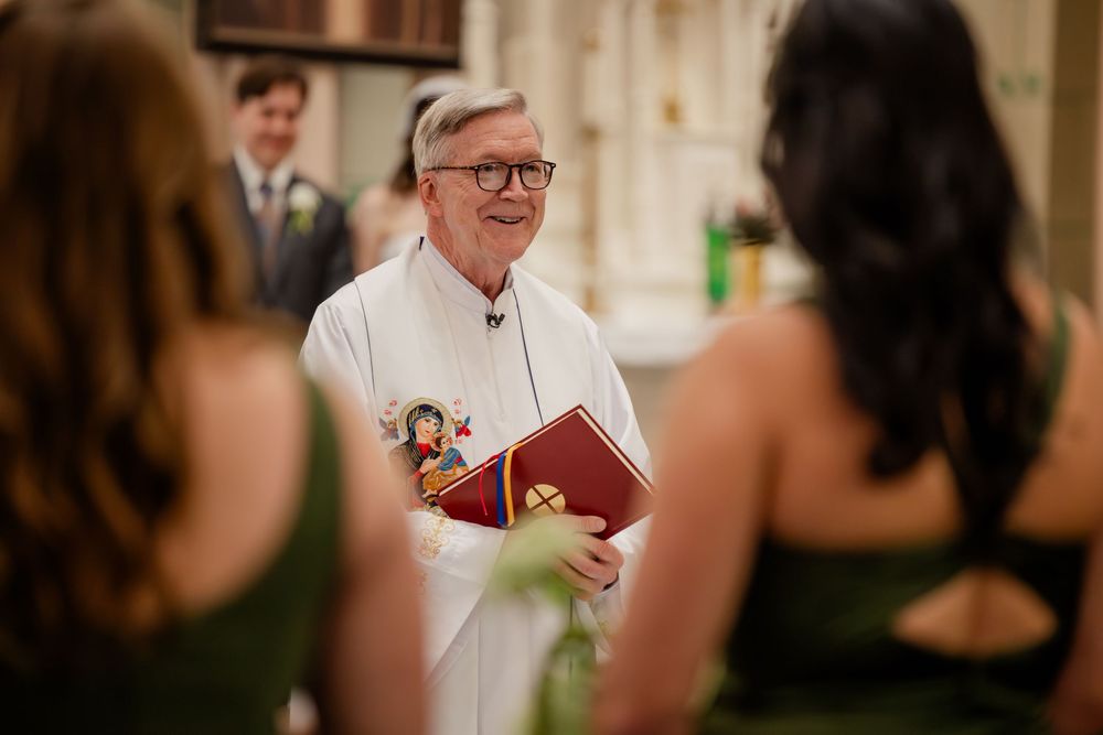 A priest in white robes and glasses speaks during a Catholic church ceremony.