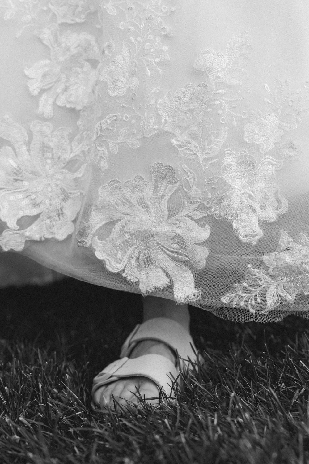 Close-up of a white lace wedding dress hem and sandal on grass in black and white.