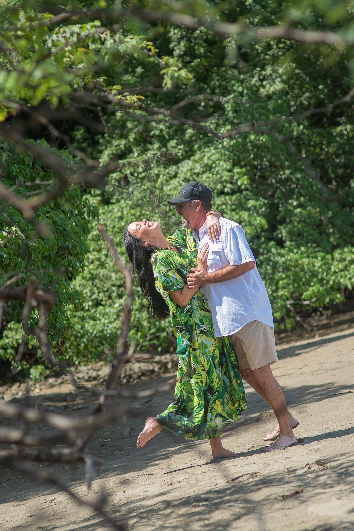 Couple in tropical attire enjoys a romantic moment on a scenic beach with lush greenery and ocean waves.