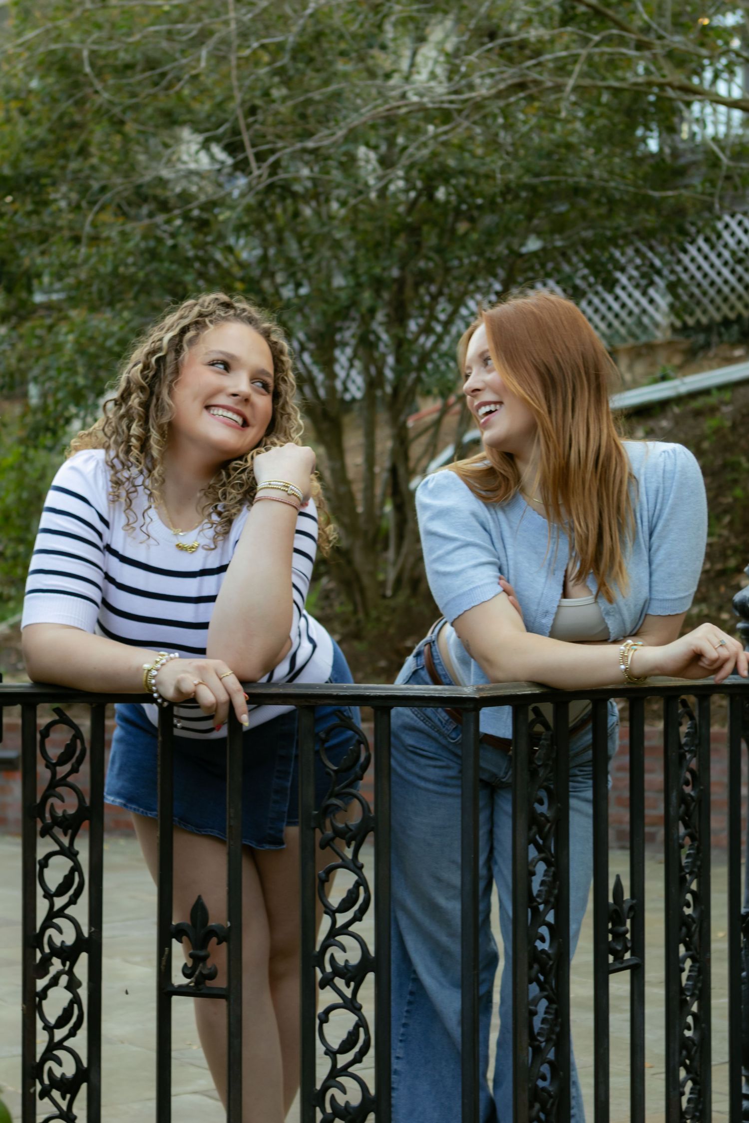 Two friends in casual attire share a lighthearted moment while leaning on a decorative iron fence.