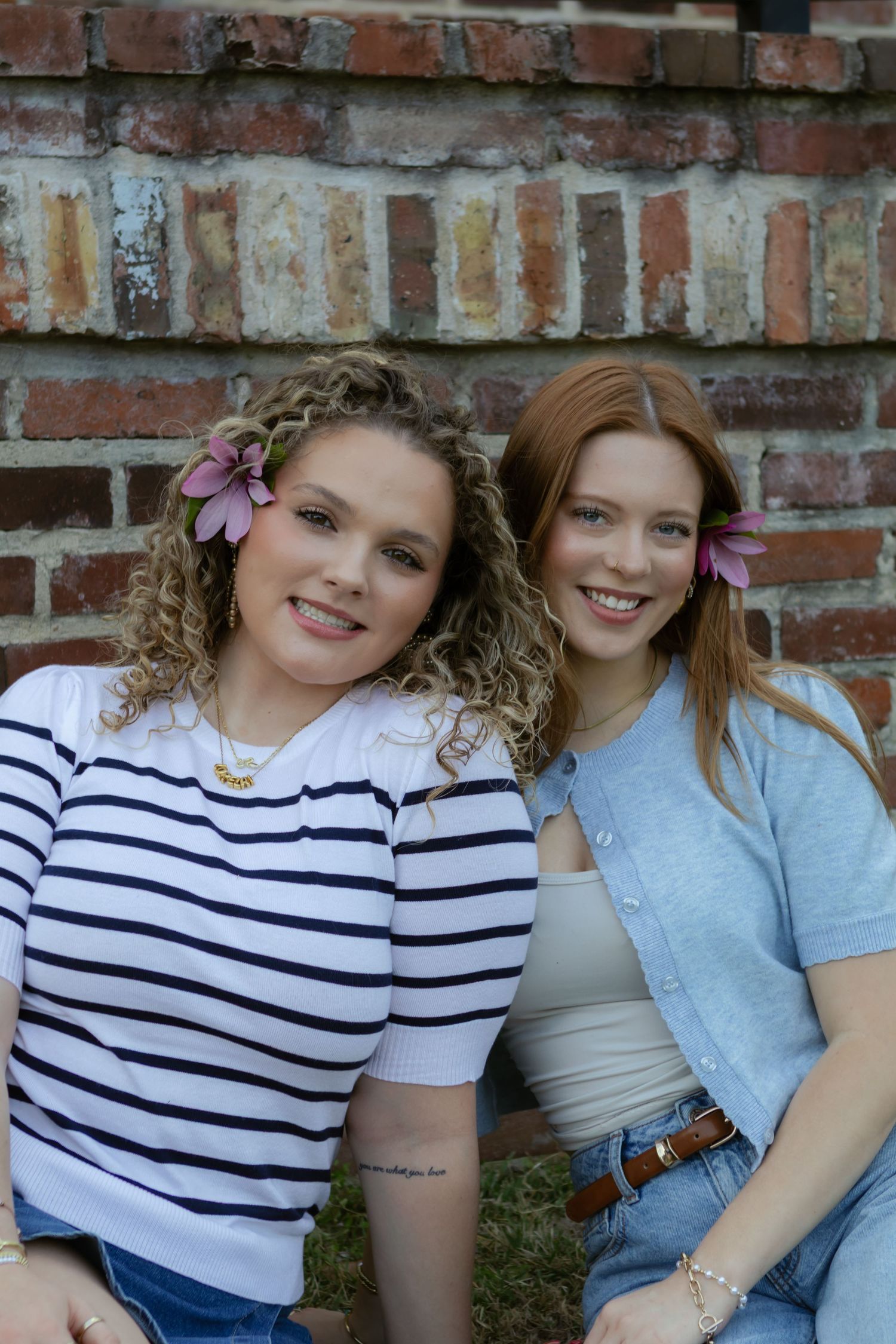 Two friends pose together against a rustic brick wall background wearing casual spring outfits and floral accessories.