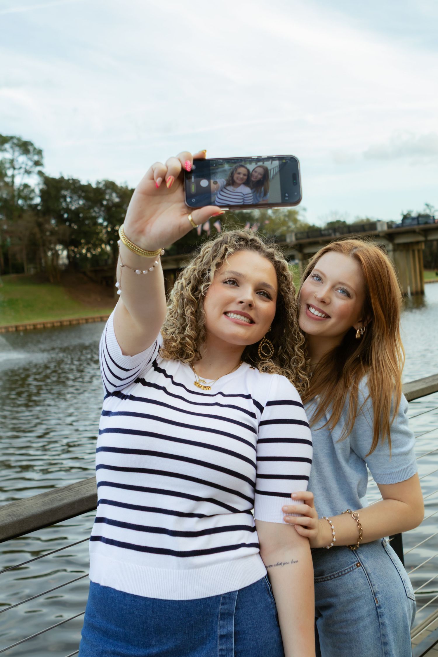 Two friends taking a selfie together by a lake on a cloudy day, one wearing stripes and one in denim.