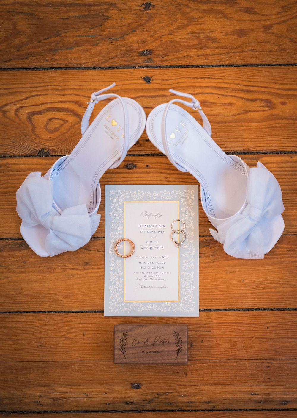 White bow heeled wedding shoes with gold rings and elegant wedding invitation on rustic wood.