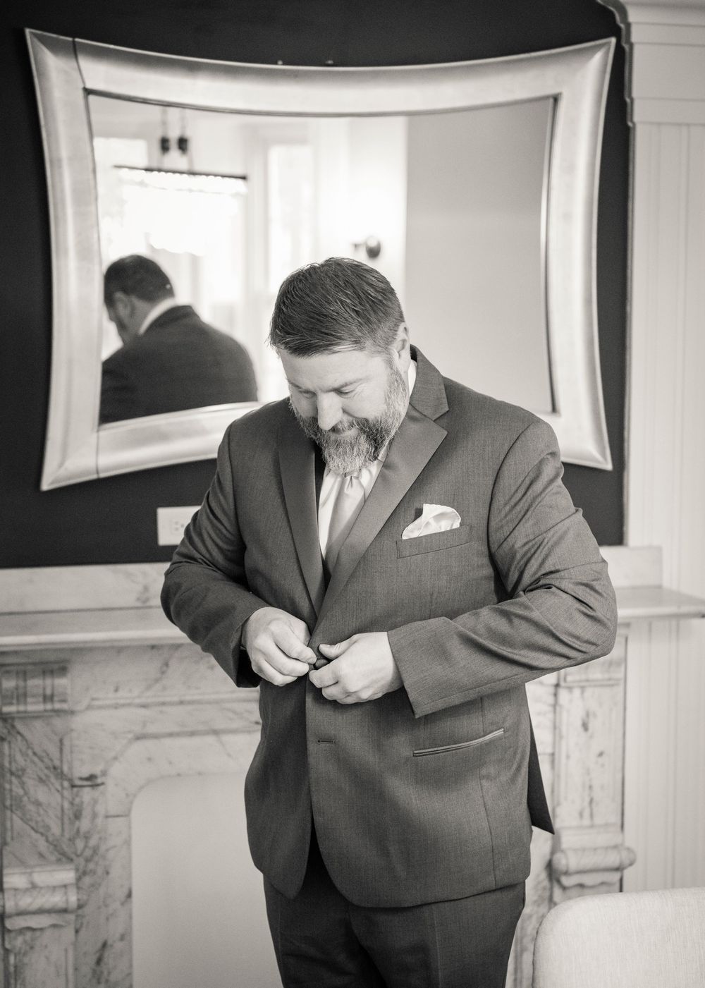 Man in gray suit buttoning jacket before mirror in black and white wedding photo.