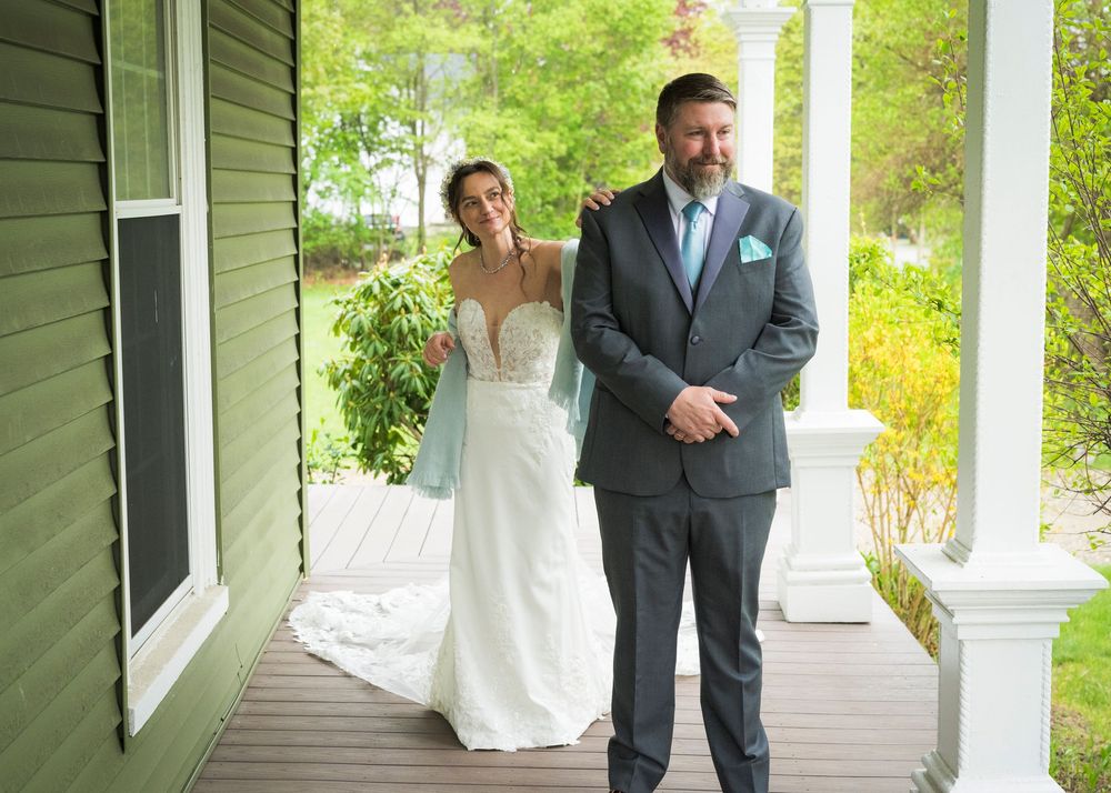Bride in white gown surprises groom in gray suit during first look on a porch at outdoor wedding.