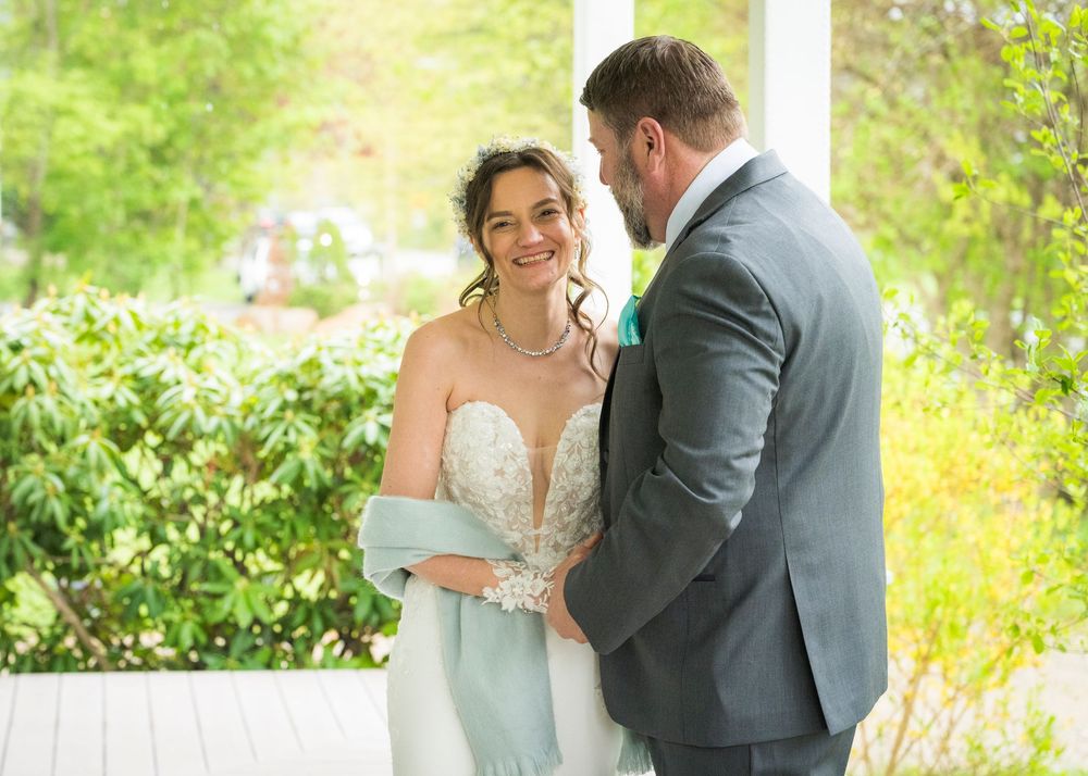 Bride and groom share a tender moment on a porch, bride in lace gown with blue wrap, groom in gray suit.
