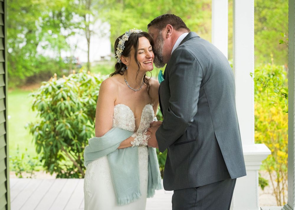 Bride and groom sharing a tender moment on a porch, bride in white gown with blue shawl, groom in gray suit.