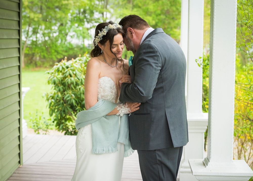 Bride and groom share an intimate moment on a porch, she in a lace gown with blue wrap, he in a gray suit.