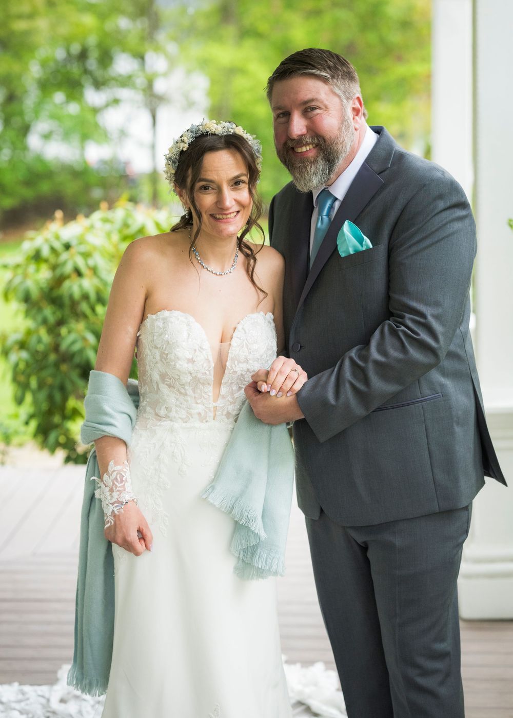 Smiling bride in lace gown with sage wrap and groom in gray suit holding hands at wedding.