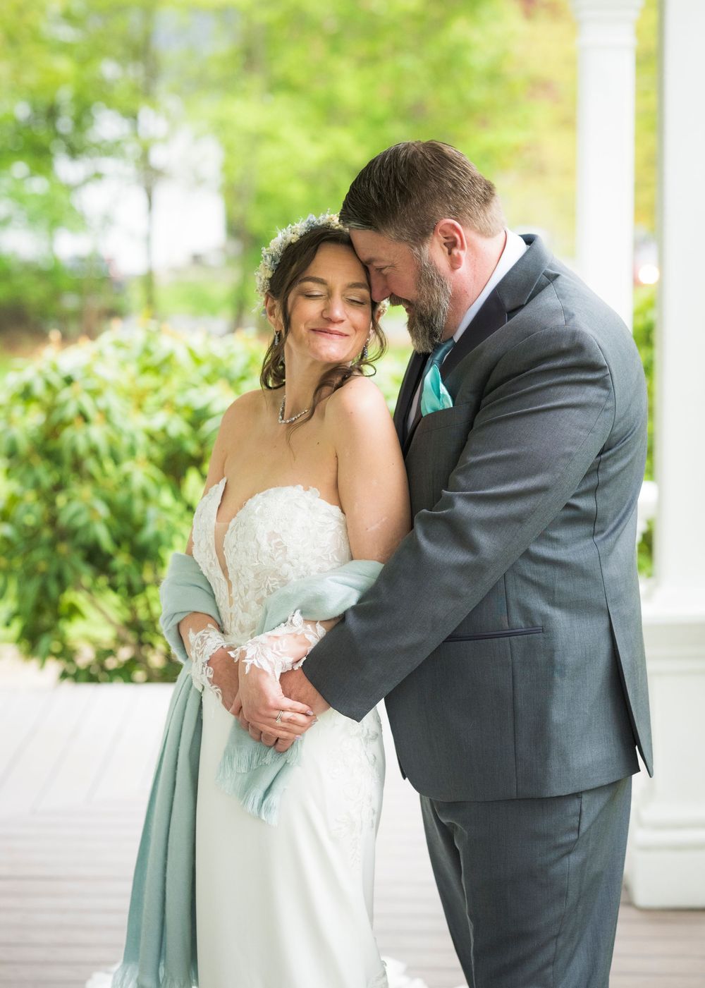Groom tenderly embracing bride in lace wedding gown with sage shawl on outdoor porch.