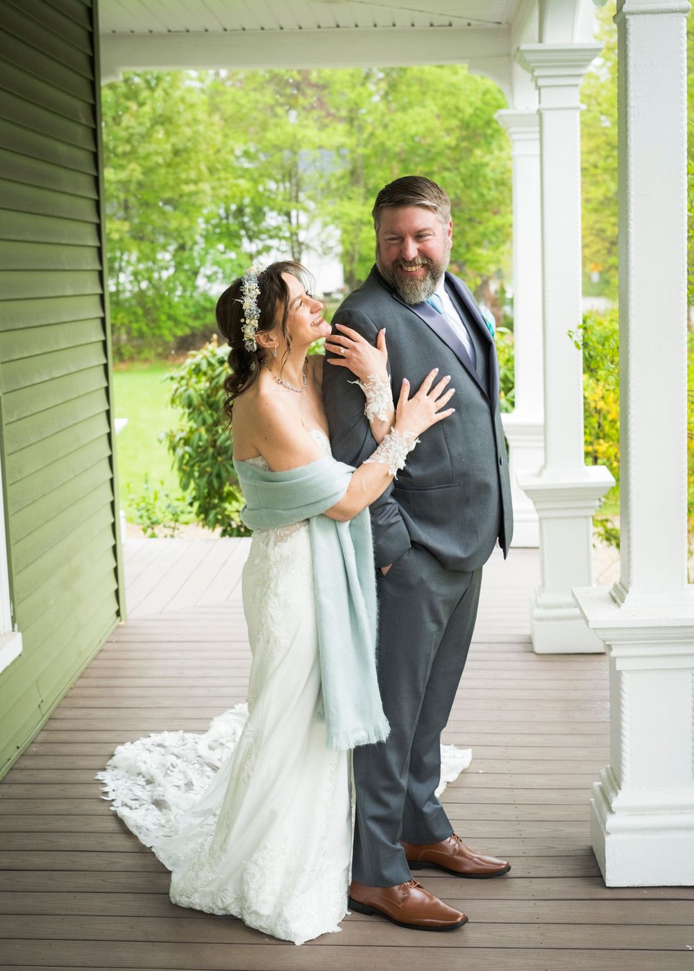 Bride in white gown with blue wrap laughs with bearded groom in gray suit on a porch.