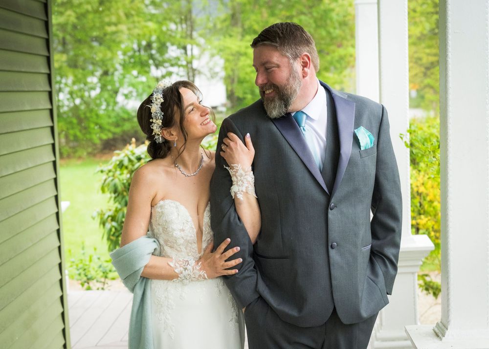 A bride and groom share a tender moment on a porch, she in a lace gown with blue wrap, he in a gray suit.