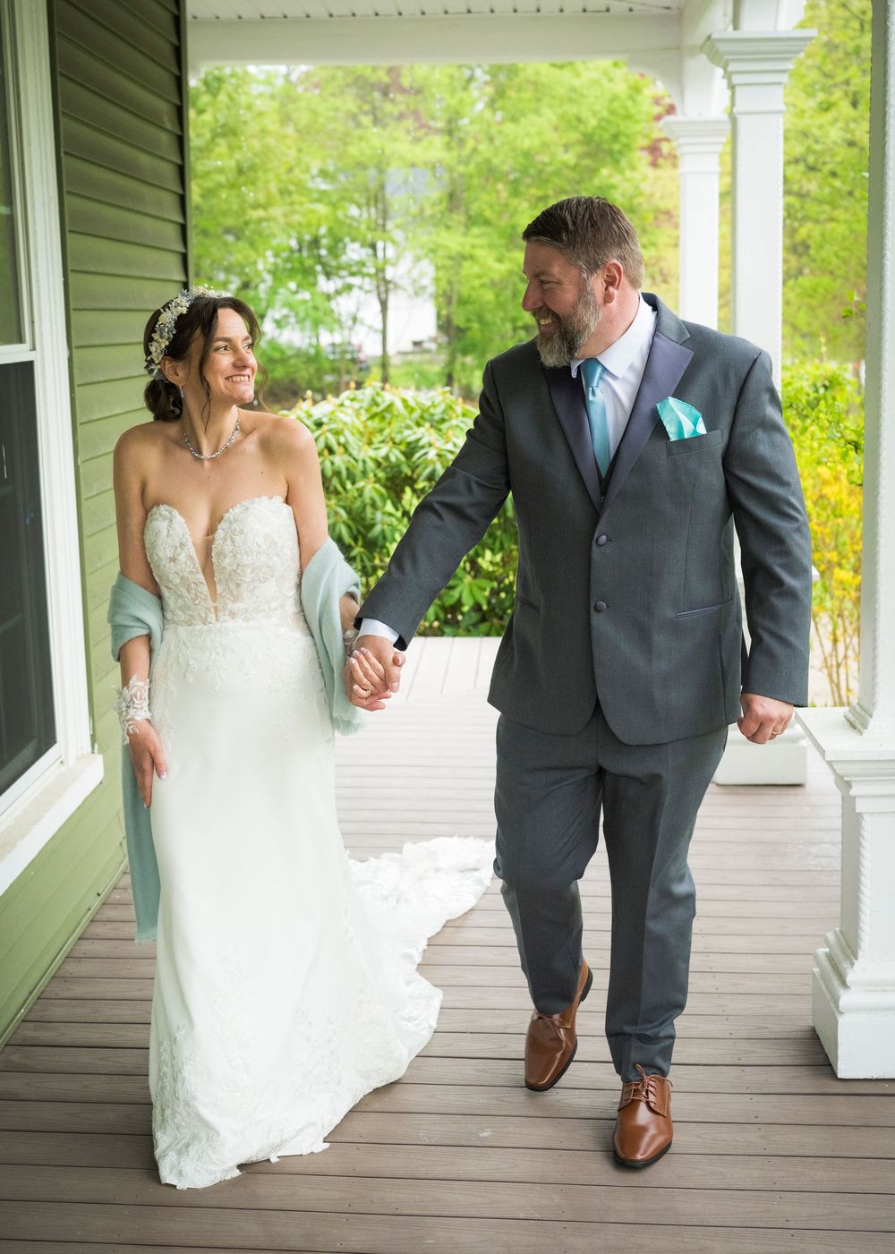 Bride and groom holding hands walking on a porch, bride in lace gown with teal wrap, groom in gray suit.