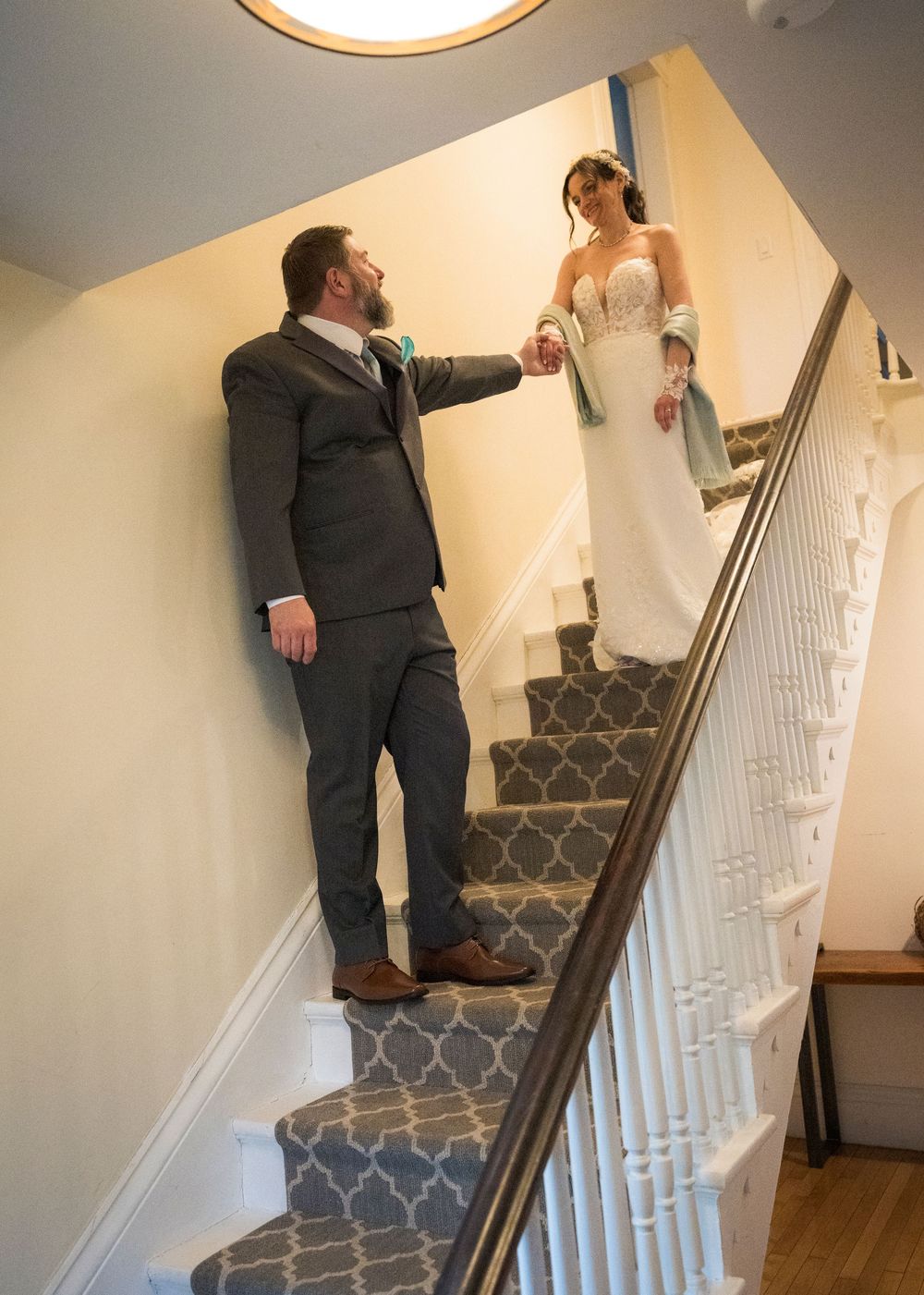 Groom holding bride's hand on elegant staircase, bride in lace gown with teal wrap, groom in gray suit.