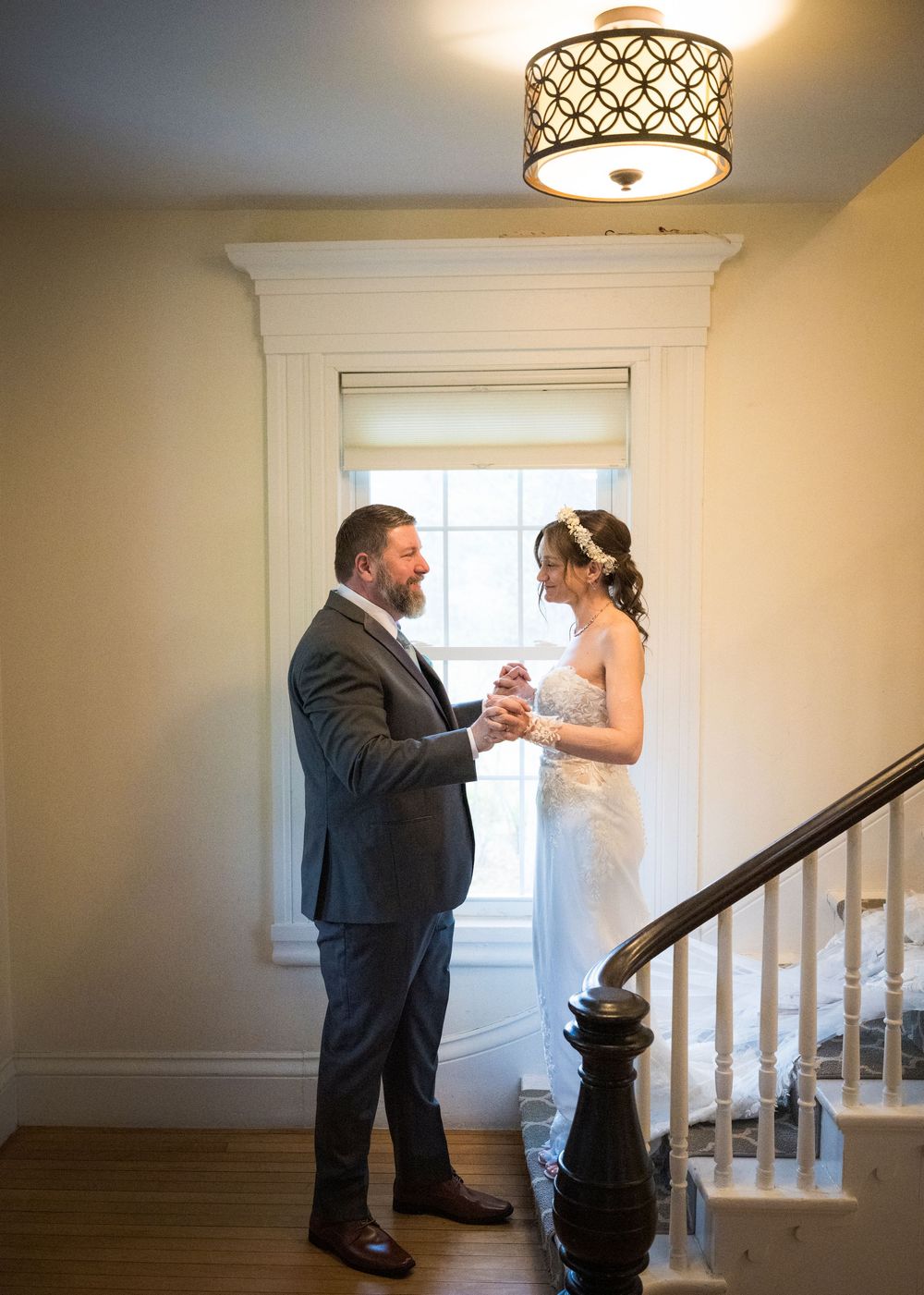 Bride and groom share a tender moment holding hands near a staircase in an elegant white interior.