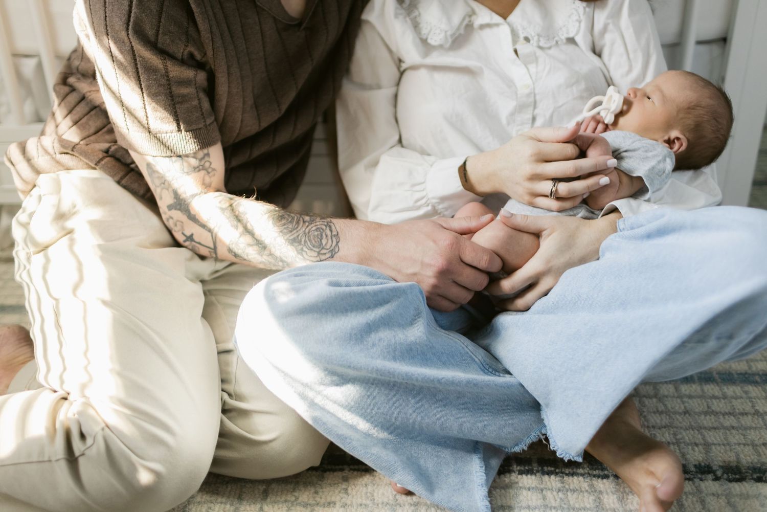 Father holds his newborn's tiny leg as the mother hold him on her lap while sitting on rug in baby nursery. Chelmsford newborn photographer.