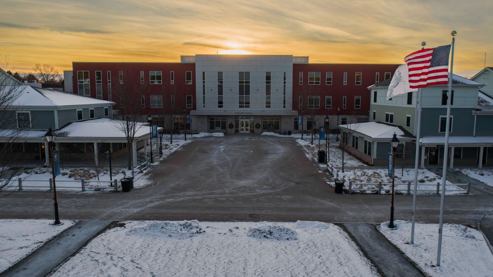 Snowy campus buildings and American flag at sunset with orange sky.
