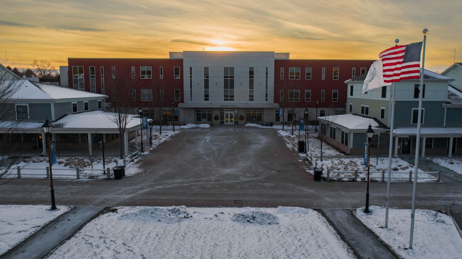 Snowy campus buildings and American flag at sunset with orange sky.
