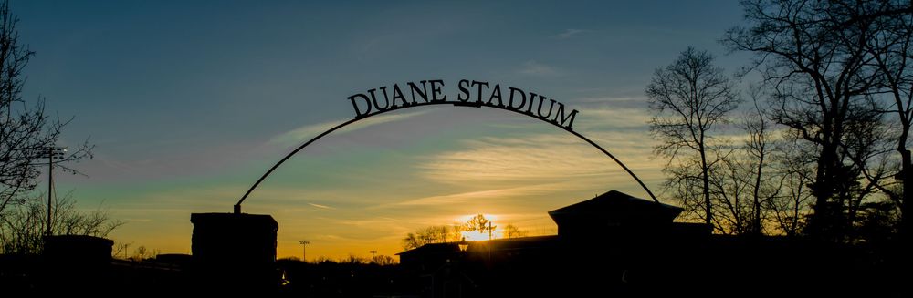 Silhouette of Duane Stadium entrance arch against colorful sunset sky.