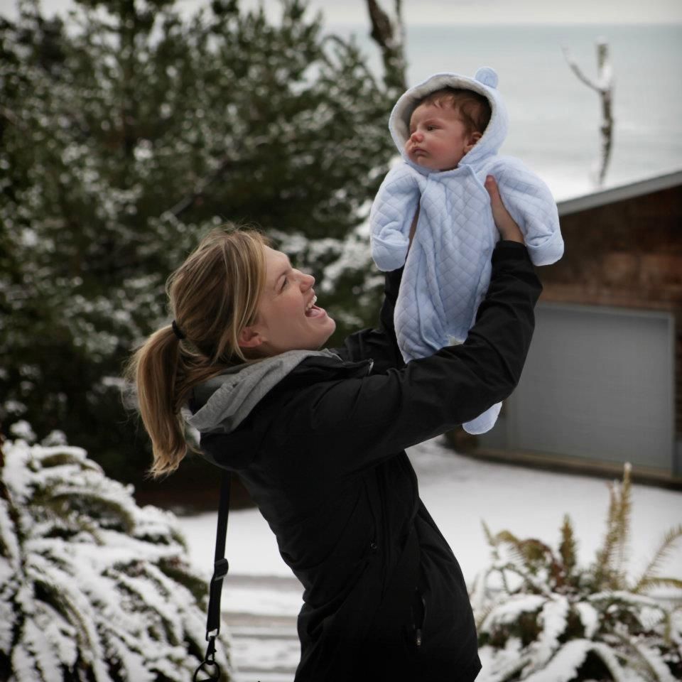Woman in black jacket lifting baby in blue snowsuit outdoors in snowy winter setting.
