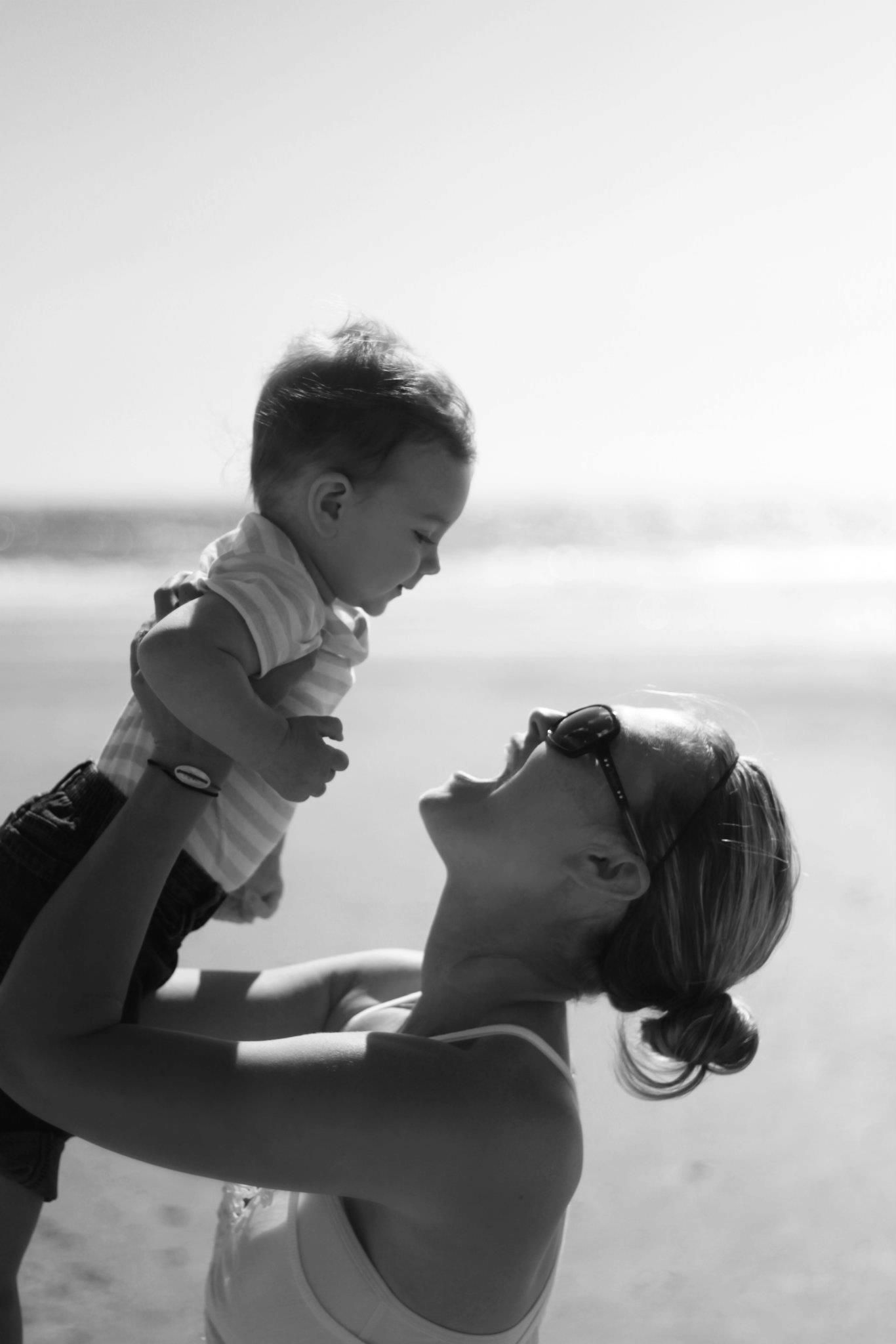 Black and white photo of mother lifting smiling toddler at sunny beach.
