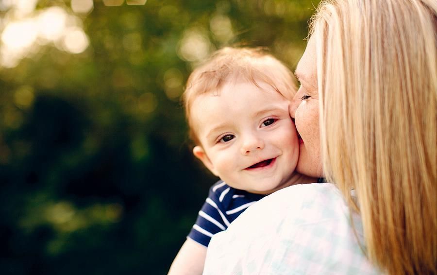Mother kissing smiling baby boy on cheek outdoors with green bokeh background.