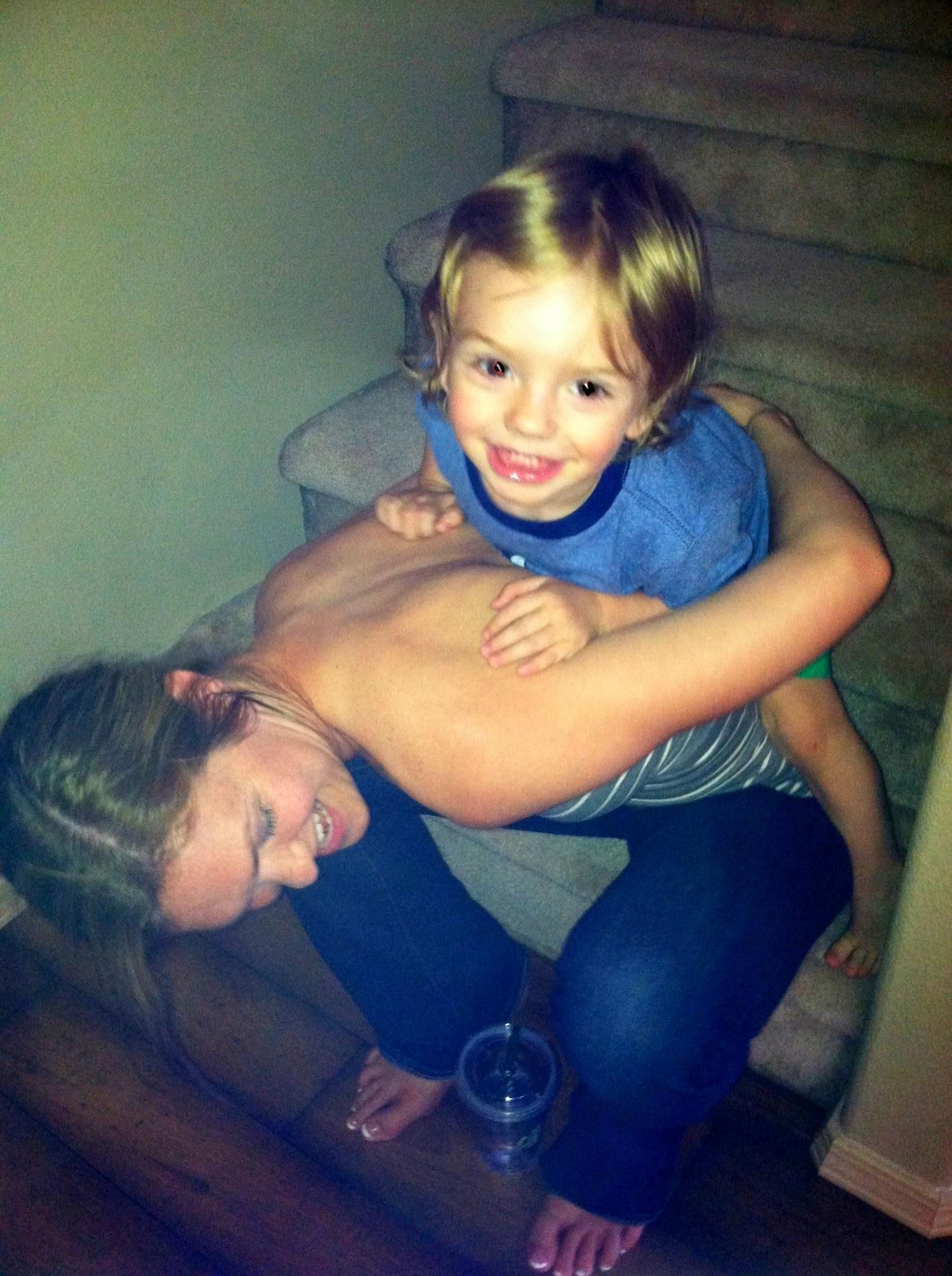 Young child playfully climbing on mothers back near staircase indoors.