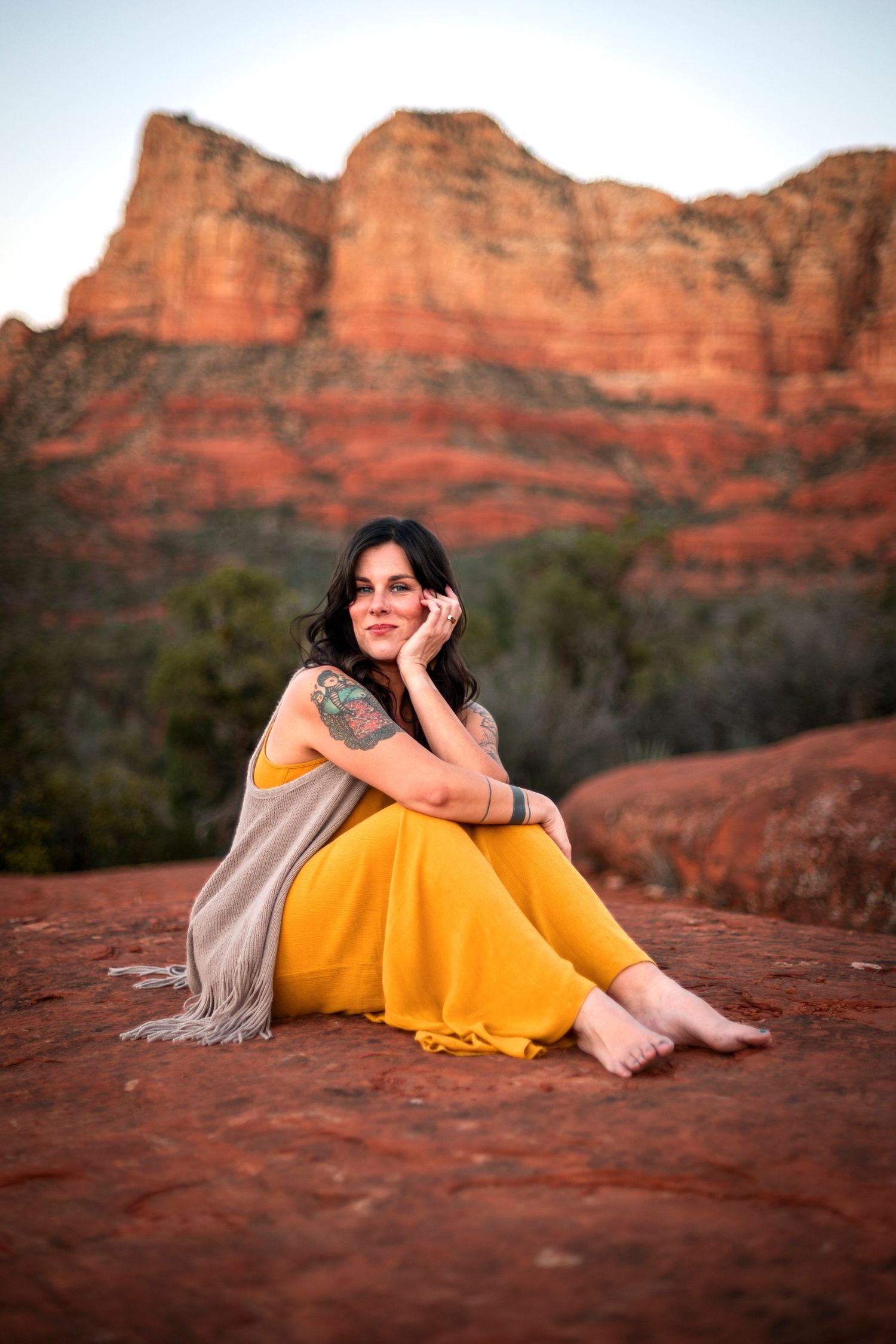 Woman in yellow outfit sits barefoot on red rocks in Sedona, Arizona, with stunning red rock formations behind her.