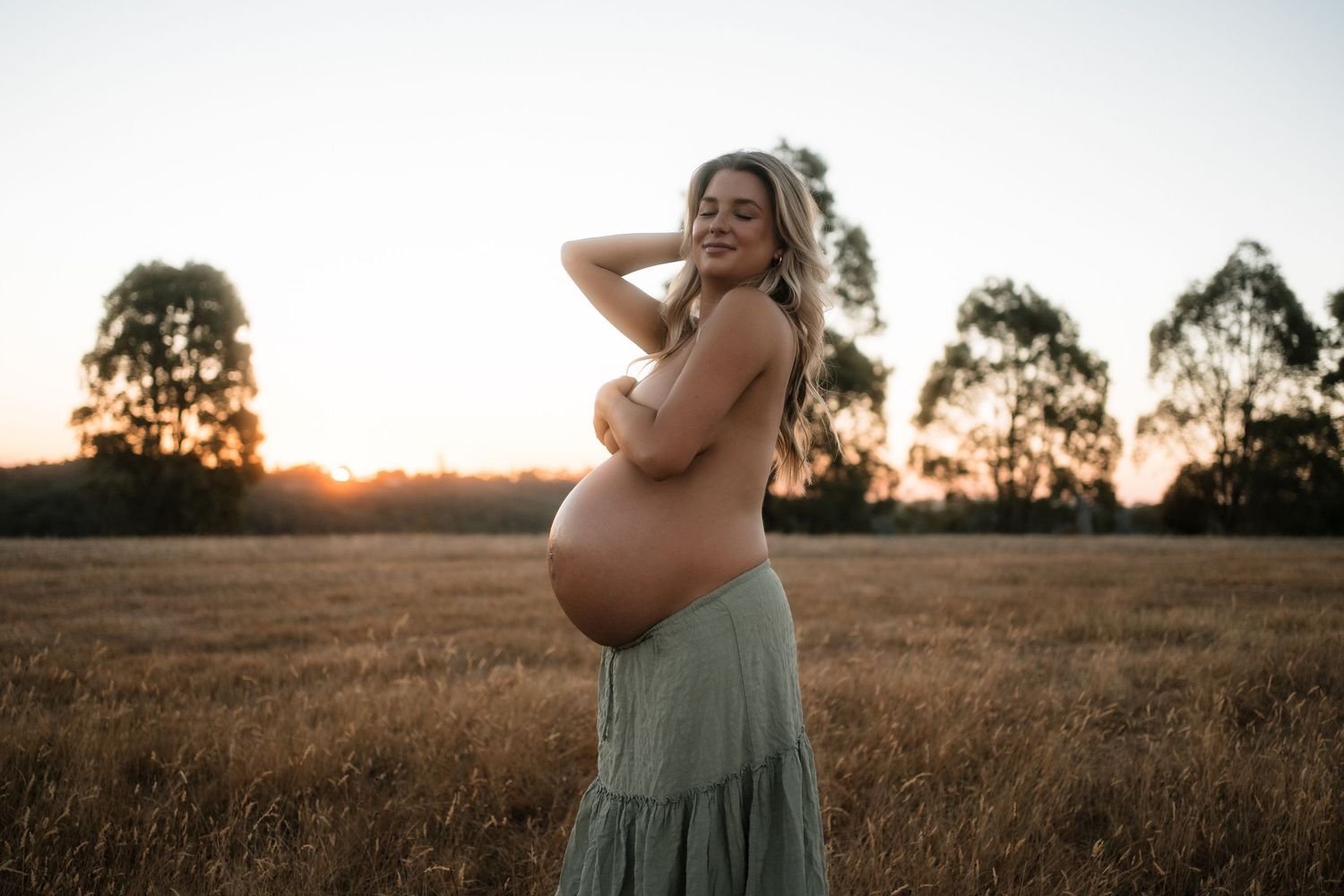 A scenic outdoor maternity photoshoot in a golden wheat field at sunset with rolling hills in the background.