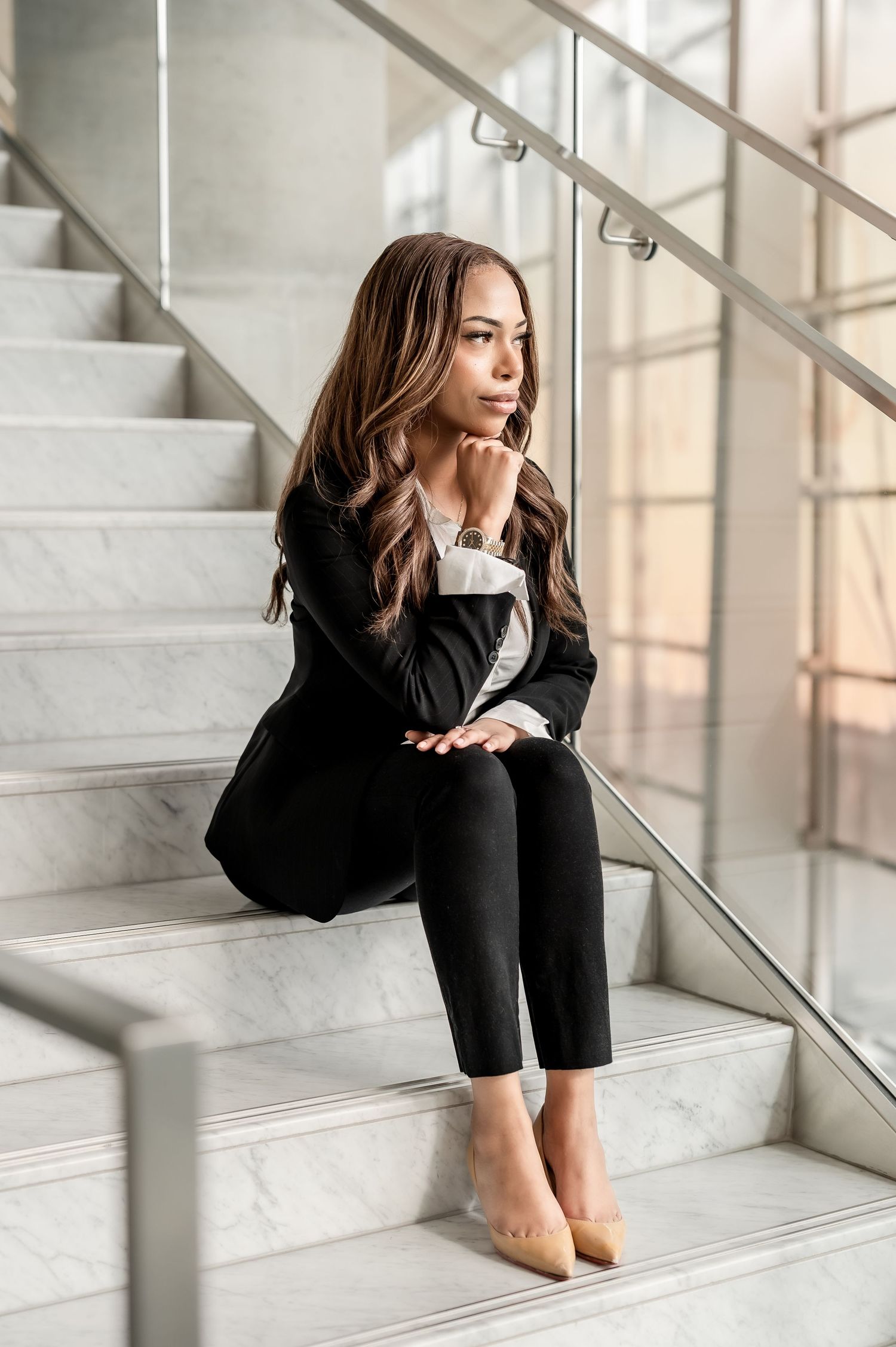 A woman in business attire sits thoughtfully on marble stairs near large windows with natural light streaming in.