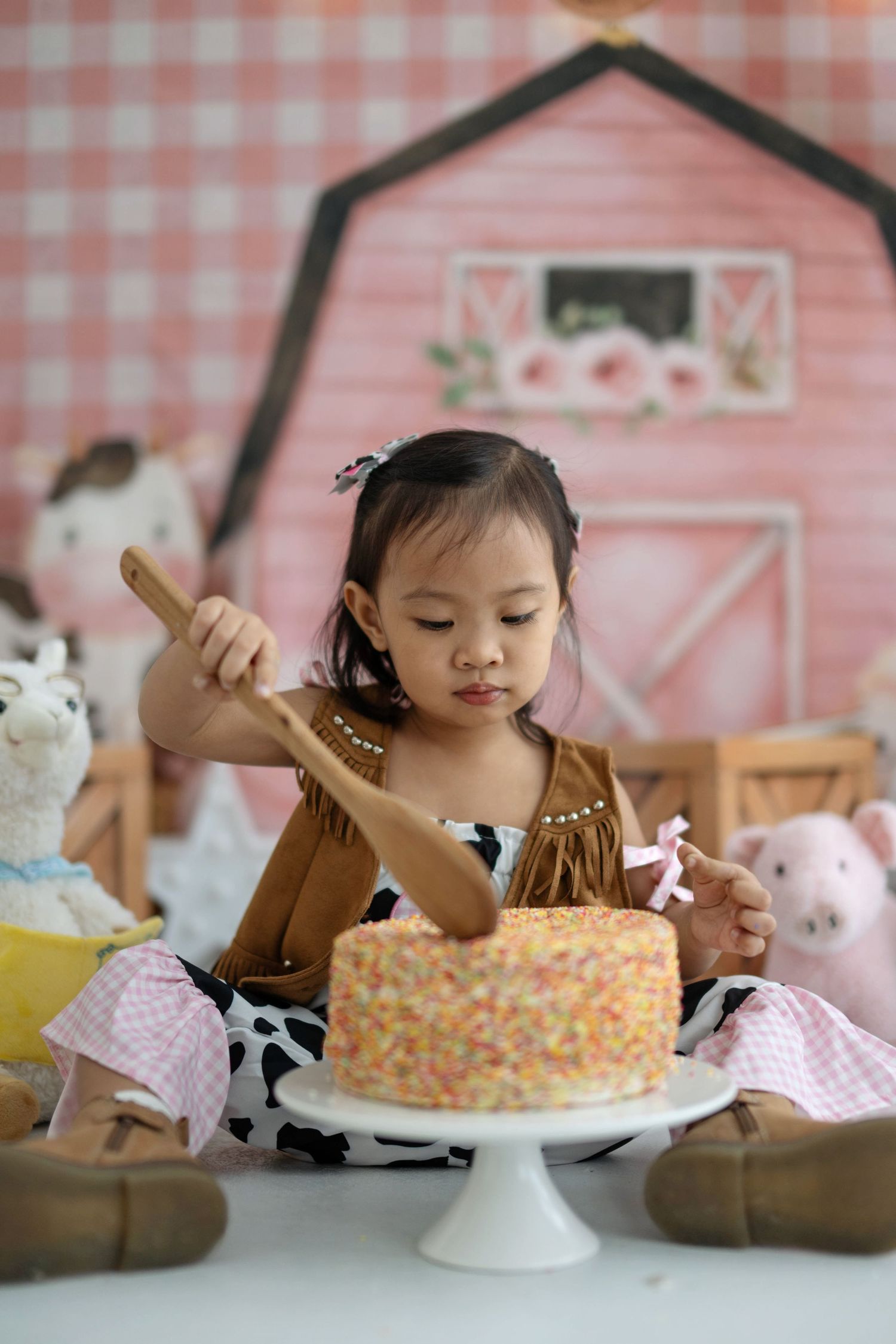 A festive cake smash setup featuring a sprinkle-covered cake on a white pedestal against a pink barn backdrop.
