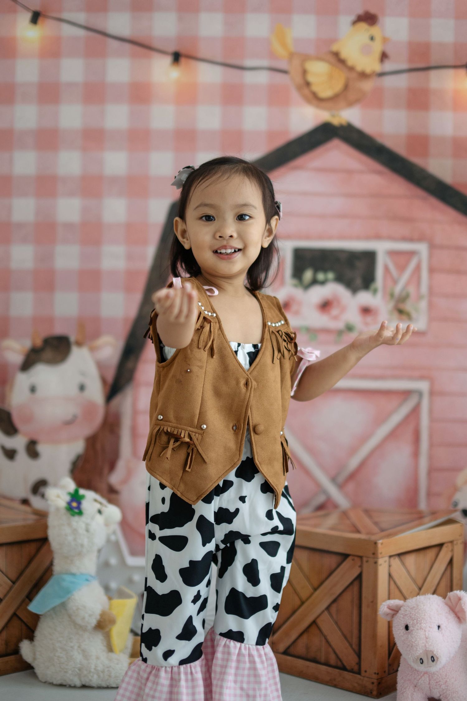 A child in a brown vest and cow print pants poses in front of a pink gingham backdrop with plush farm animals.