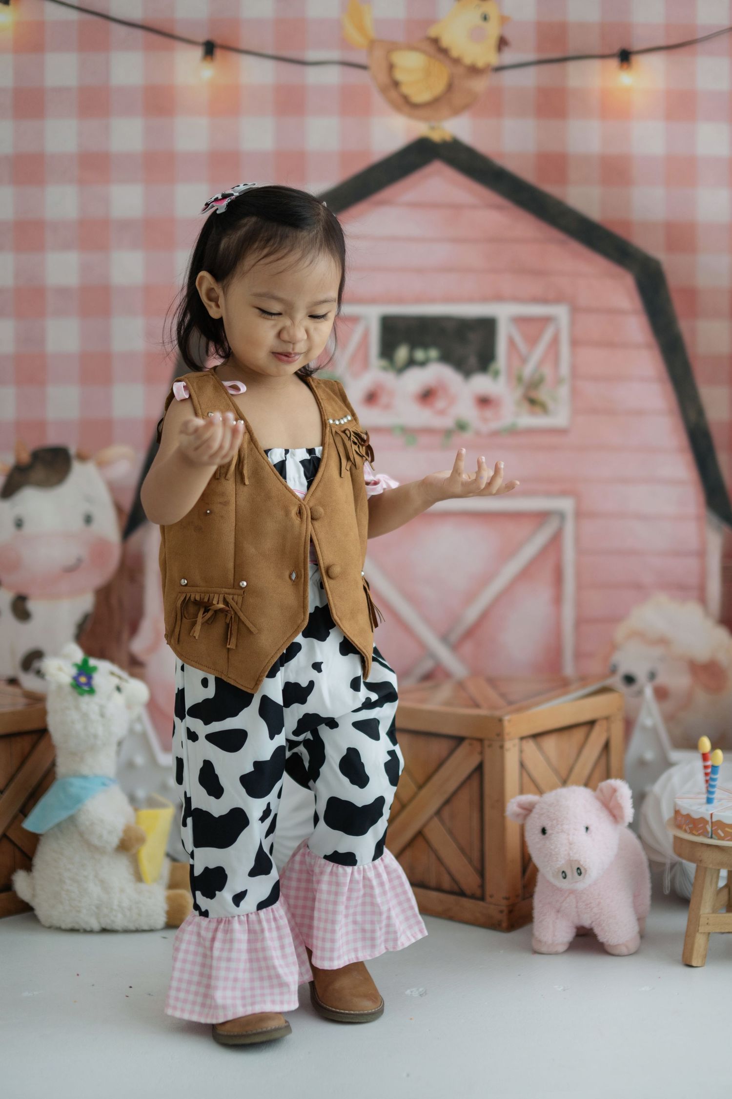 A child in a cow print outfit and brown vest plays with farm-themed toys and stuffed animals against a pink backdrop.